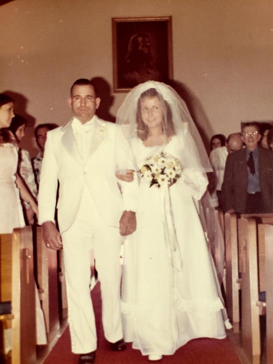 A bride in white walks with her groom, surrounded by guests in a church.