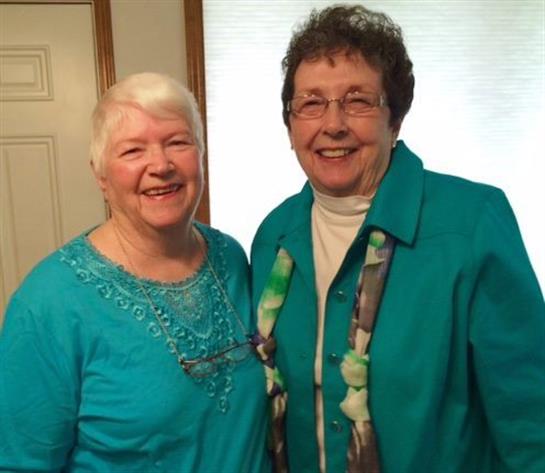 Two cheerful older women dressed in turquoise stand together, smiling warmly at home.