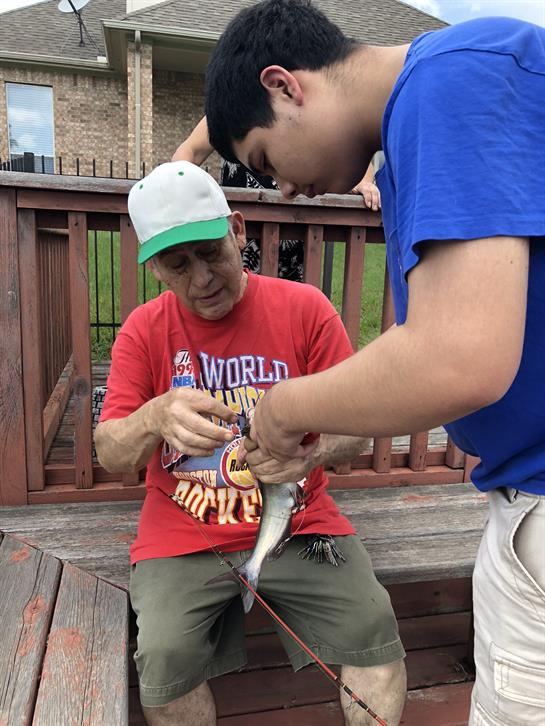 A man shows a young boy how to properly hold and prepare a fish at a lakeside deck.