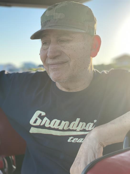 A cheerful older man wearing a cap and casual shirt sits outside, basking in warm sunset light.