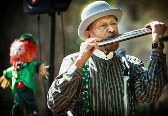 An elderly musician plays the flute at a lively outdoor gathering with a puppet show.