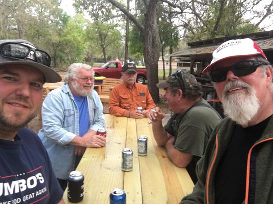 Five friends chat animatedly over drinks at a rustic picnic table in nature.