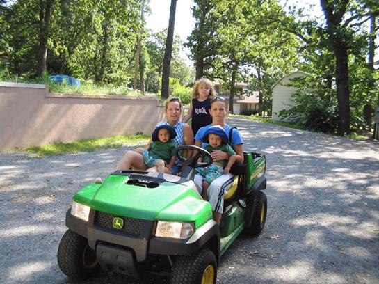 A family of four takes a joyful ride in a green vehicle along a gravel road surrounded by trees.