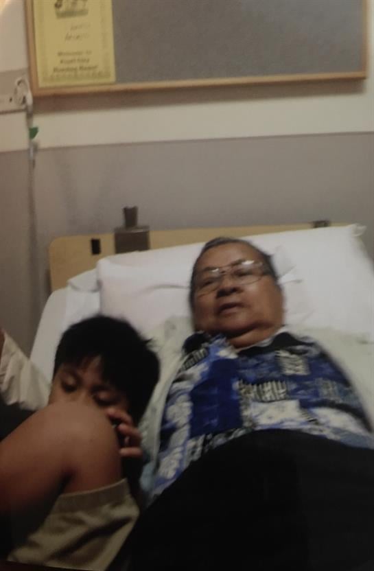 An elderly woman lies in hospital bed while a young child sits beside her, showing affection.