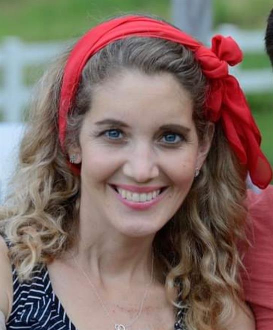 A woman with curly hair and a red headband smiles in a sunny outdoor setting.