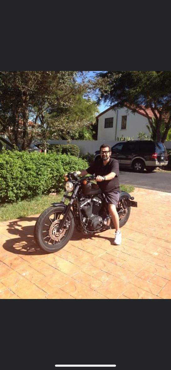 A man enjoys riding a motorcycle in a driveway surrounded by greenery under clear blue skies.