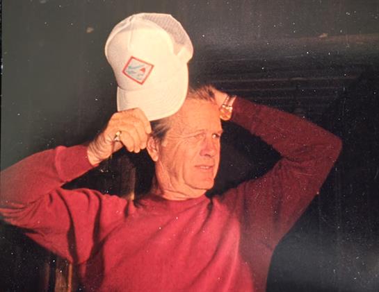 A man in a red sweater is playfully adjusting a white hat with a goofy expression while indoors.