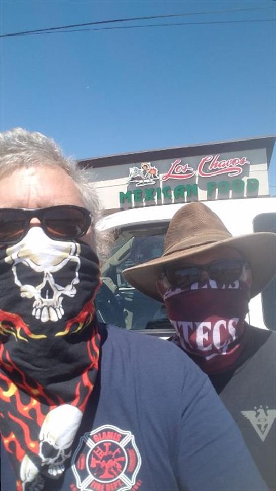 Two men with sunglasses wearing masks stand outdoors in front of a restaurant under clear skies.