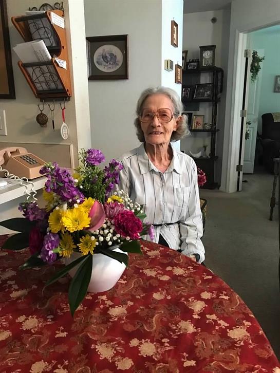An elderly woman smiles warmly while seated at a round table adorned with a vibrant flower bouquet.