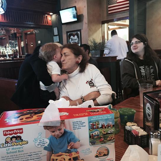 A young child kisses their mother while they sit at a restaurant table with family enjoying a meal.