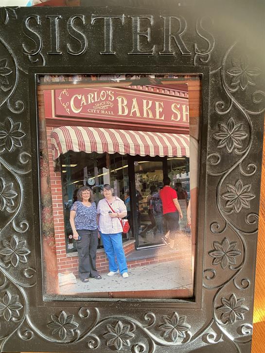 Two women smile in front of a bakery with a bright red storefront and awning.