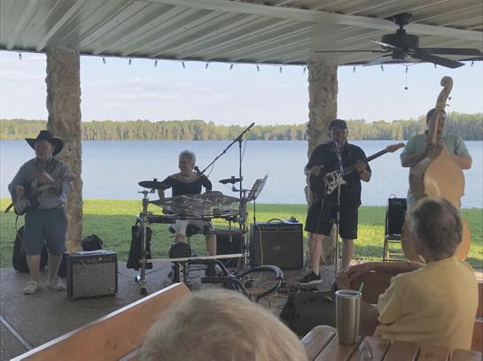 Musicians entertain an audience at a lakeside venue on a beautiful sunny day while seated.