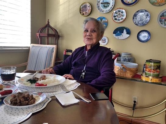 A smiling elderly person sits at a wooden table with a plate of food, enjoying a cozy meal at home.