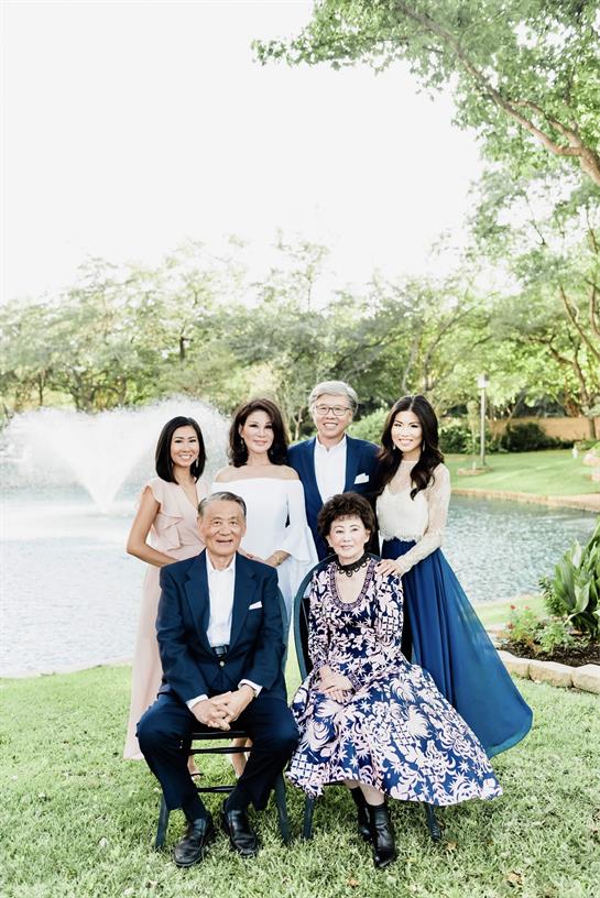 Family members pose happily by a lake, dressed in elegant attire surrounded by greenery.