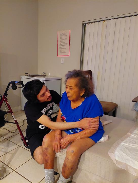 A caregiver shows affection to an elderly woman while sitting on a bed in a warm, homey environment.