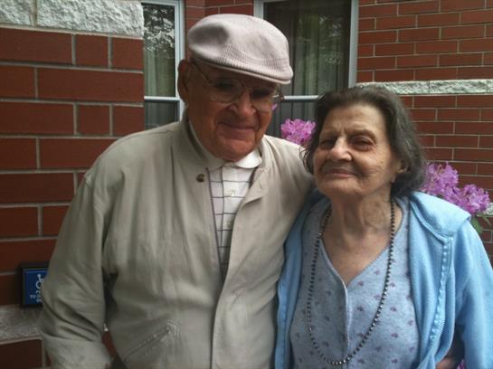 An elderly couple stands together, smiling warmly, enjoying time outside their home.