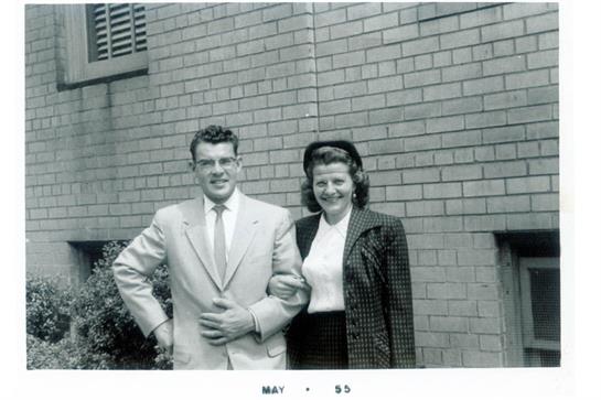 Man in suit and woman in stylish outfit pose together near brick wall in bright May weather.