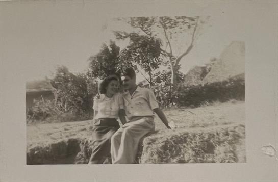 Two people smile and sit closely together on a grassy area under the shade of trees.