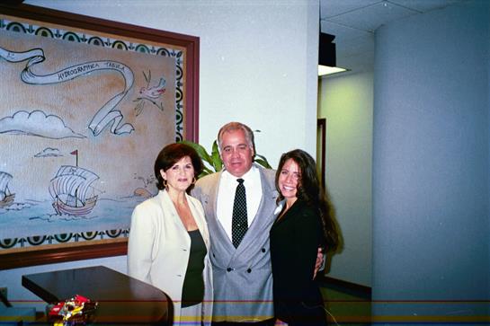 Three professionals stand together in an office, smiling and dressed in business attire.