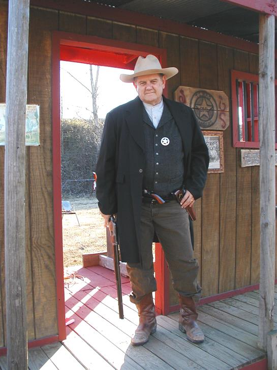 A confident sheriff poses at a western-style doorway, showcasing his traditional attire and firearm.