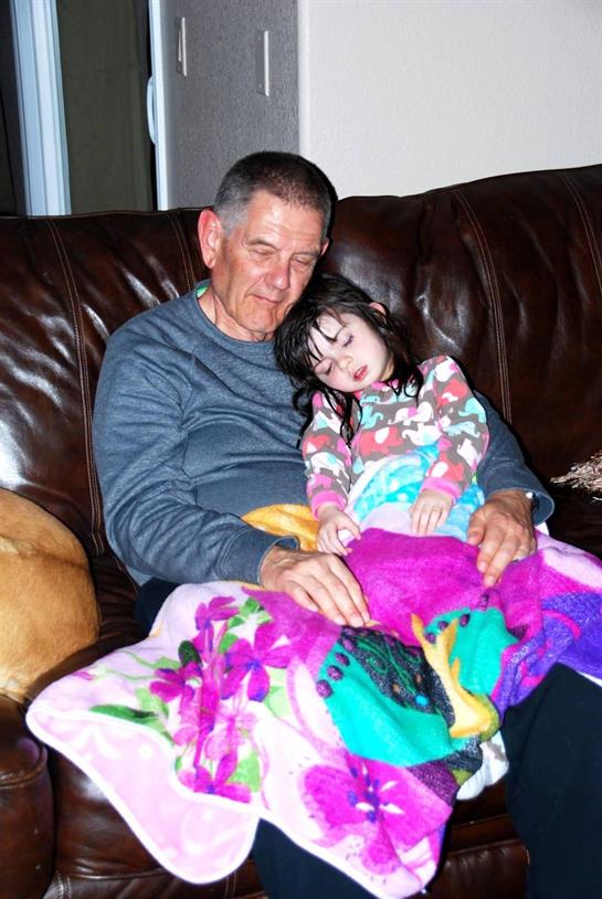 A grandfather shares a quiet moment with his sleepy granddaughter on the couch, dog nearby.
