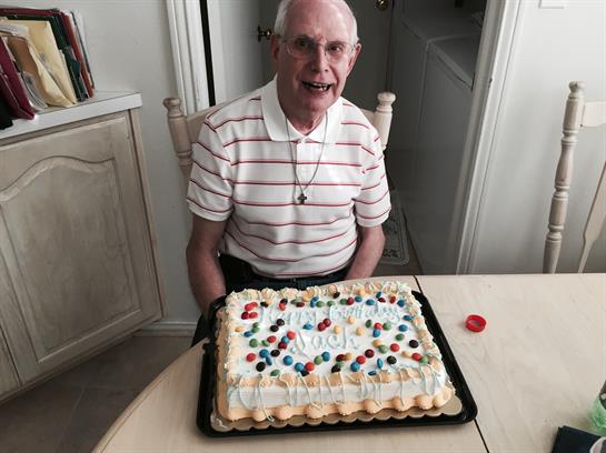 An elderly man happily sits at a table with a brightly decorated birthday cake.