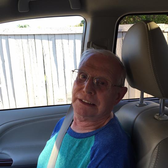 Elderly man smiles while seated in a car, a sunny afternoon setting outside the window.