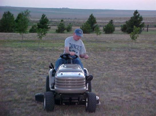 A man is cutting grass with a riding lawn mower in a field during sunset, surrounded by trees.