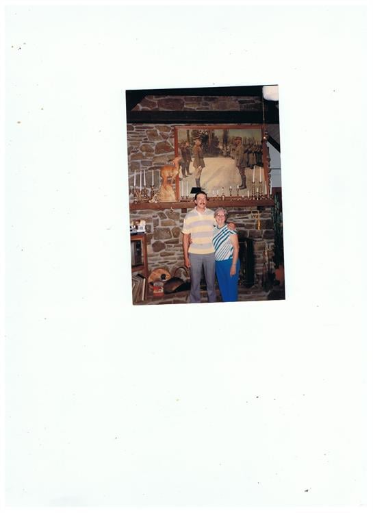Two individuals stand together, smiling in front of a rustic stone fireplace adorned with decor.