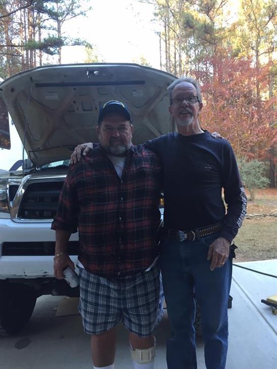 Friends stand together near a truck that has its hood raised in a wooded area.