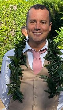 A joyful man wears a floral garland and light attire, smiling during a daytime celebration outdoors.