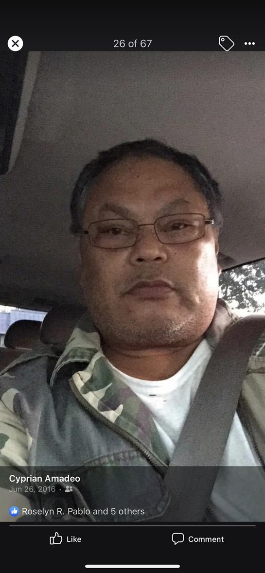 A man in glasses wears a camo jacket, sitting seriously in a car's driver's seat.