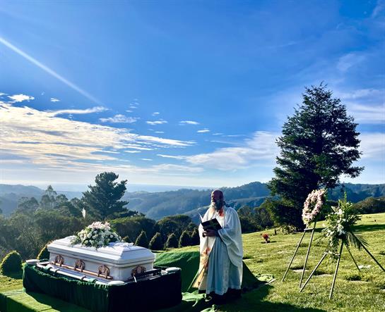 A solemn ceremony is held at a peaceful cemetery site surrounded by mountains and a clear blue sky.