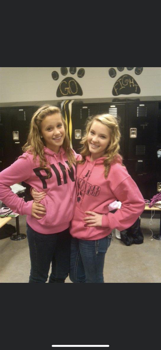 Two girls pose happily in pink sweatshirts, enjoying their time in a school locker room.