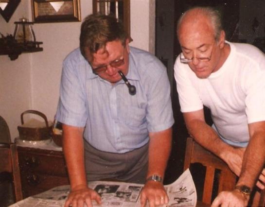 Two elderly men study vintage newspapers on a table, sharing memories in a cozy indoor setting.