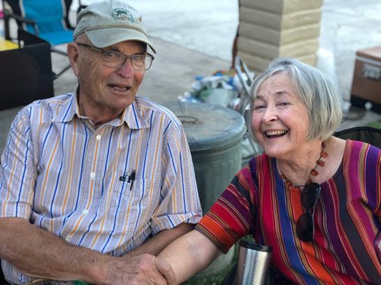 An elderly couple is smiling and holding hands while sitting in a lively outdoor setting.