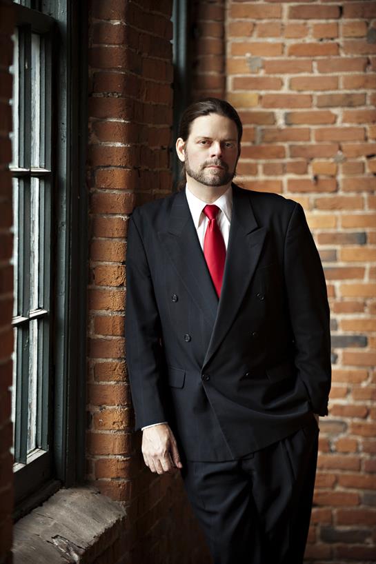 A man dressed in a formal suit stands near a brick wall, exuding confidence and style.