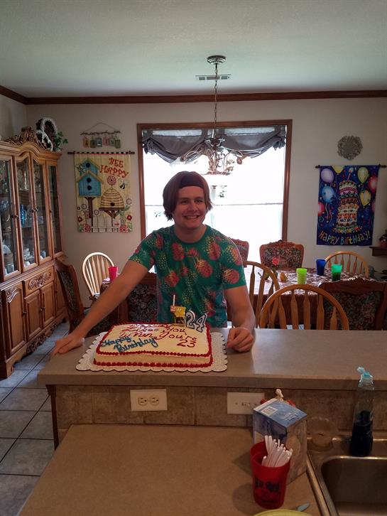 A smiling individual stands by a birthday cake in a decorated kitchen filled with cheerful items.