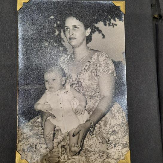 A woman sits outdoors in a floral dress, holding a baby in light clothing, under a tree.