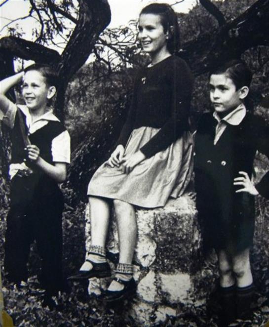 Three children stand and sit on a stone, enjoying a moment outdoors with nature around them.