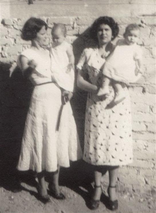 Two women dressed in vintage dresses hold babies while enjoying a sunny day outdoors.