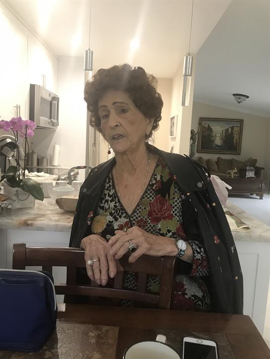 An elderly woman in a patterned outfit sits at a table engaging with someone in a cozy home.