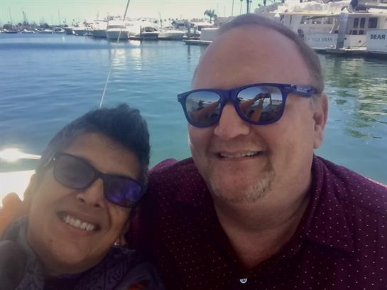 Two people smiling together, wearing sunglasses, with a beautiful marina and yachts behind them.