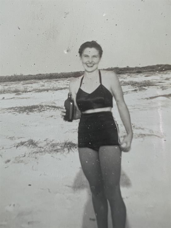 A woman smiles while standing on the beach in a retro swimsuit, enjoying her drink.