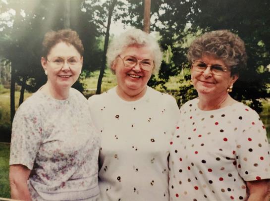 Three women joyfully stand outdoors, dressed in matching polka dot shirts, surrounded by greenery.