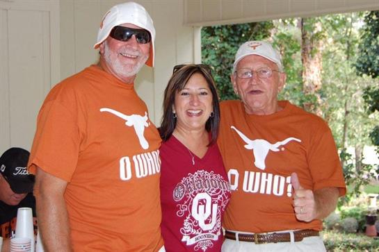 Three friends proudly pose in team shirts while celebrating a college football rivalry at home.