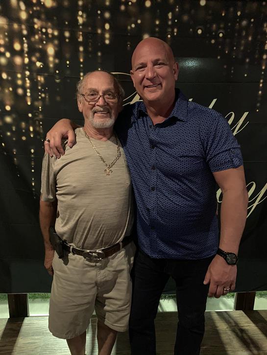Two men stand happily side by side, smiling confidently in a vibrant setting with lights.