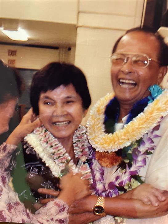 Friends joyfully embrace a couple adorned with flower leis during a festive gathering.