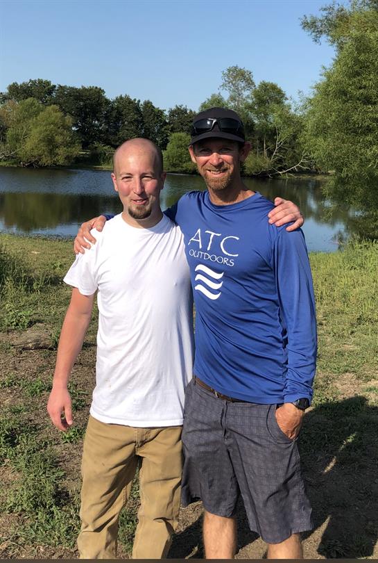 Two friends stand together by a peaceful lake, enjoying a warm day outdoors in good company.