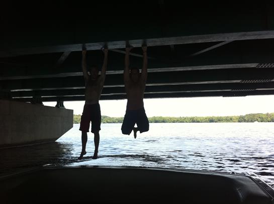 Two young individuals joyfully hanging from a bridge above the river, enjoying their time outdoors.